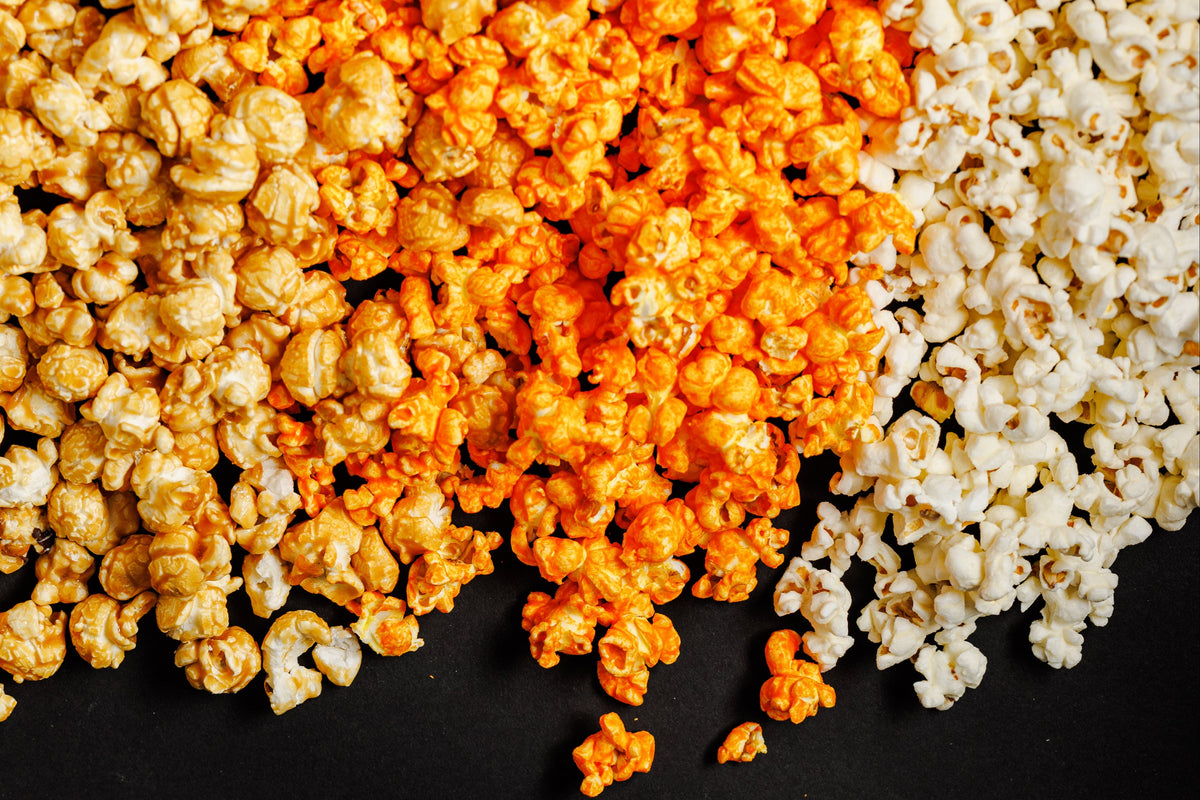 Three types of popcorn arranged in diagonal bands on a black background: plain white popcorn, orange-flavored popcorn, and golden caramel popcorn.