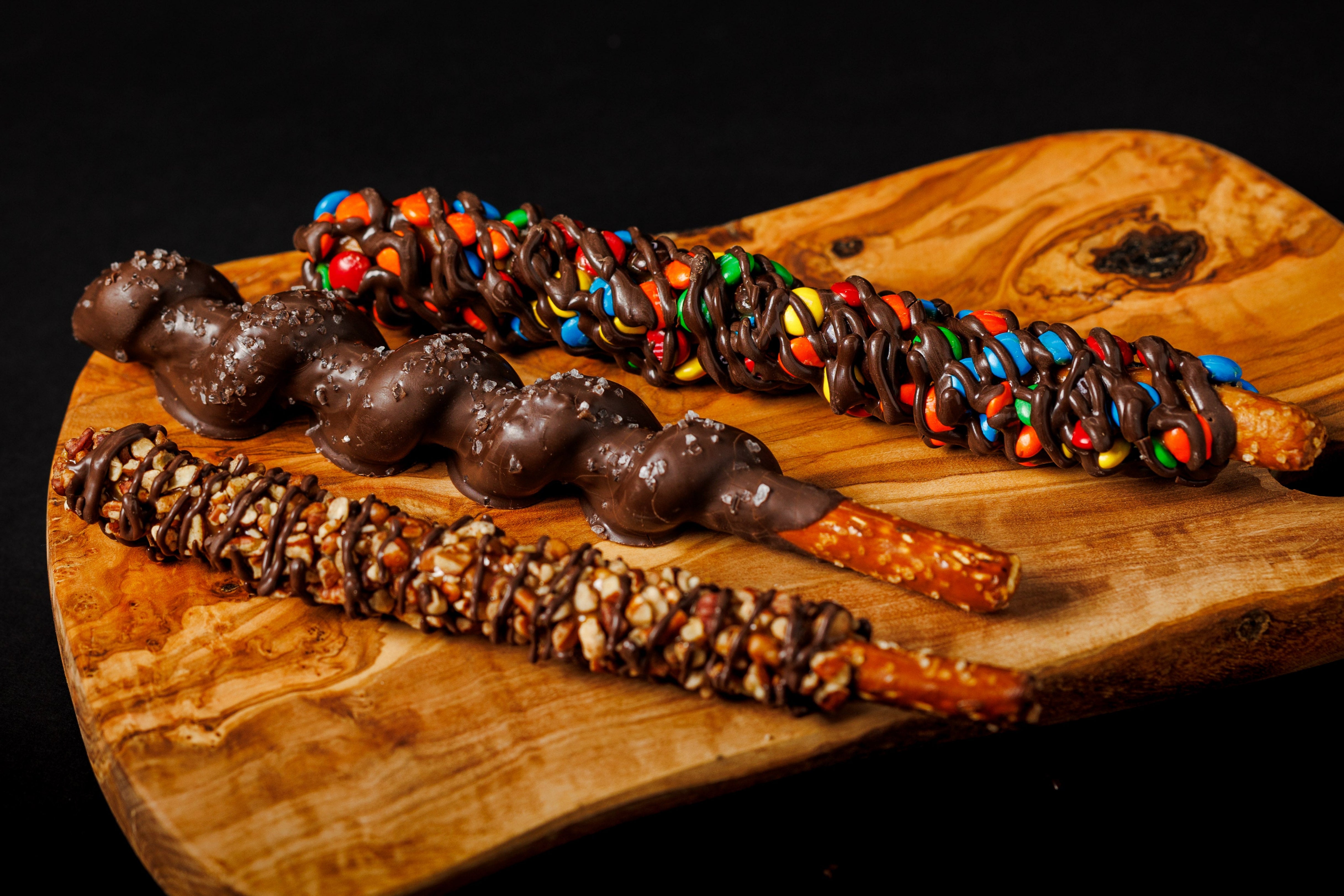 A selection of gourmet pretzels with various toppings, including nuts and candies, displayed on a wooden board.