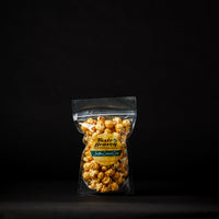 A clear plastic stand-up pouch filled with caramel popcorn, standing on a black surface against a black background. The pouch has a yellow and green label reading 'Taste of Heaven' and 'Buttery Caramel Popcorn'.