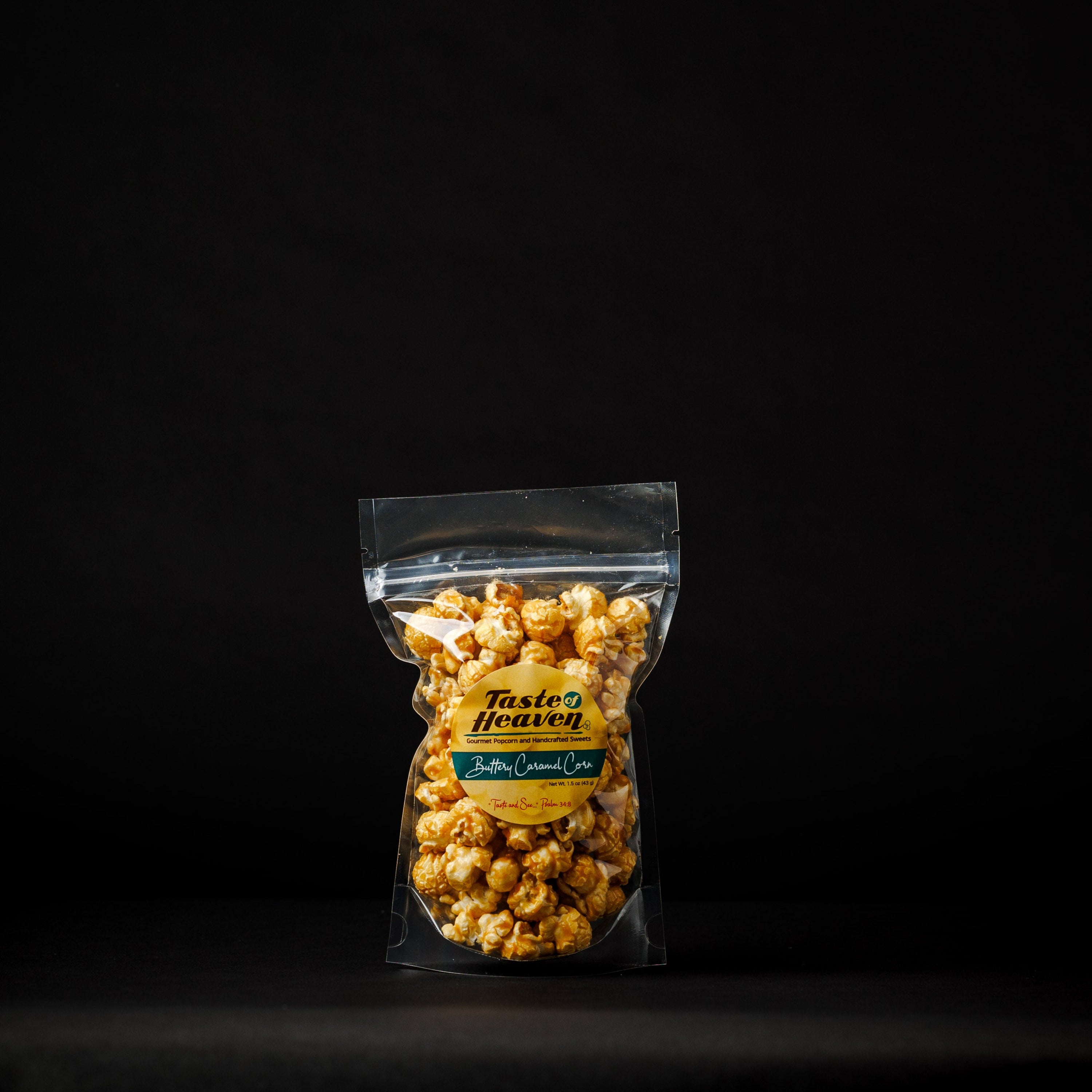 A clear plastic stand-up pouch filled with caramel popcorn, standing on a black surface against a black background. The pouch has a yellow and green label reading 'Taste of Heaven' and 'Buttery Caramel Popcorn'.