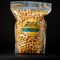 A clear plastic stand-up pouch filled with buttery caramel popcorn, featuring a round yellow and teal 'Taste of Heaven' label, set against a solid black background.
