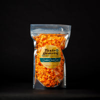 A clear plastic stand-up pouch filled with bright orange cheese-flavored popcorn, featuring a round yellow and teal label that reads 'Taste of Heaven' and 'Cheddar Cheese Corn,' photographed against a solid black background.