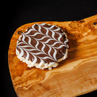 A round chocolate rice crispy treat topped with a white and dark brown swirled chocolate glaze, presented on a rustic live-edge wooden board against a solid black background.