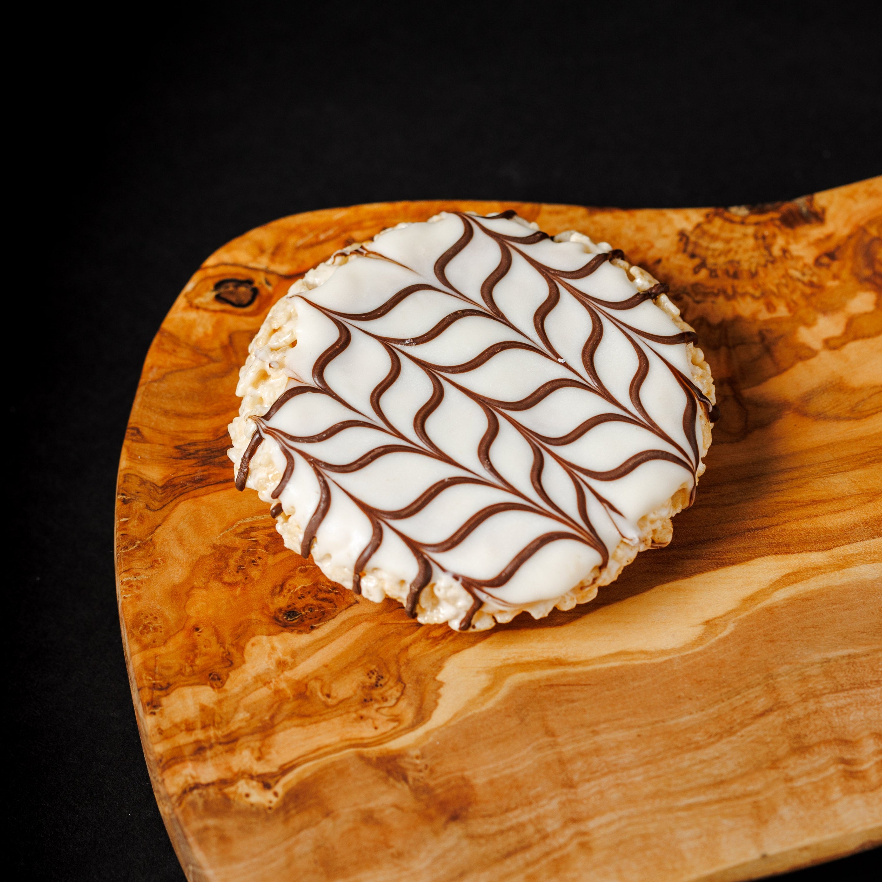 A round white rice crispy treat with brown drizzle and white chocolate, placed on a curved wooden serving board against a black background.