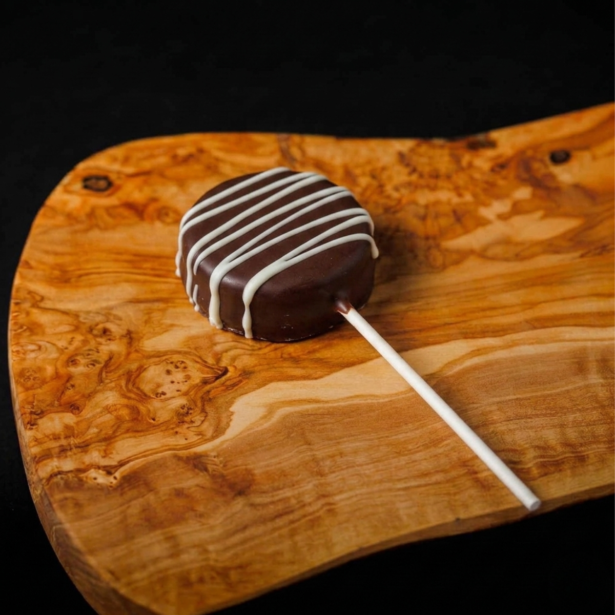 A chocolate-covered cookie with white drizzle on a white stick, placed on a curved wooden board against a black background.