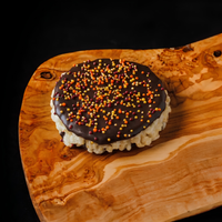 A round rice crispy treat topped with dark chocolate and colorful sprinkles, presented on a rustic olive wood serving board against a black background.