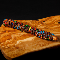 A chocolate-covered pretzel dipped in caramel and coated with M&M's candy is presented diagonally on a polished, wavy wooden board against a solid black background.