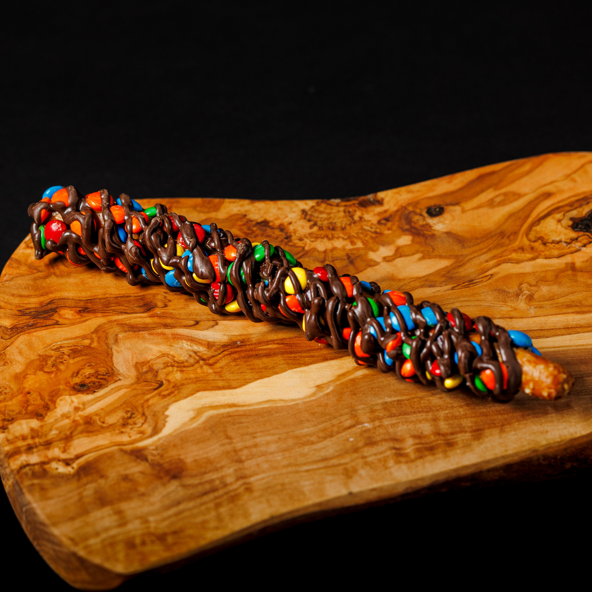 A chocolate-covered pretzel dipped in caramel and coated with M&M's candy is presented diagonally on a polished, wavy wooden board against a solid black background.