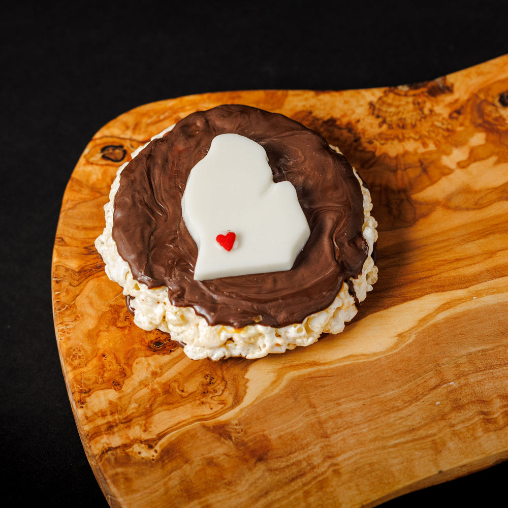 A rice crispy treat coated with caramel and dark chocolate, topped with a white chocolate Michigan mitten shape on a wooden board.