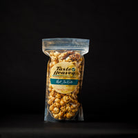 A clear plastic package of Must Be Nuts caramel corn with nuts against a dark background, branded as 'Taste of Heaven'.