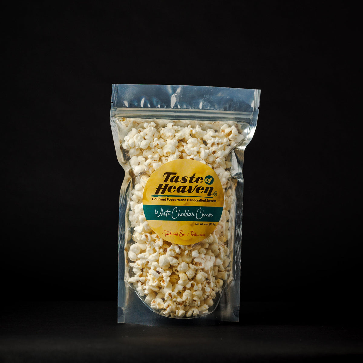A clear stand-up pouch filled with popcorn on a black background. The pouch has a yellow circular label reading 'Taste of Heaven' and 'White Cheddar Cheese' with a green band below.