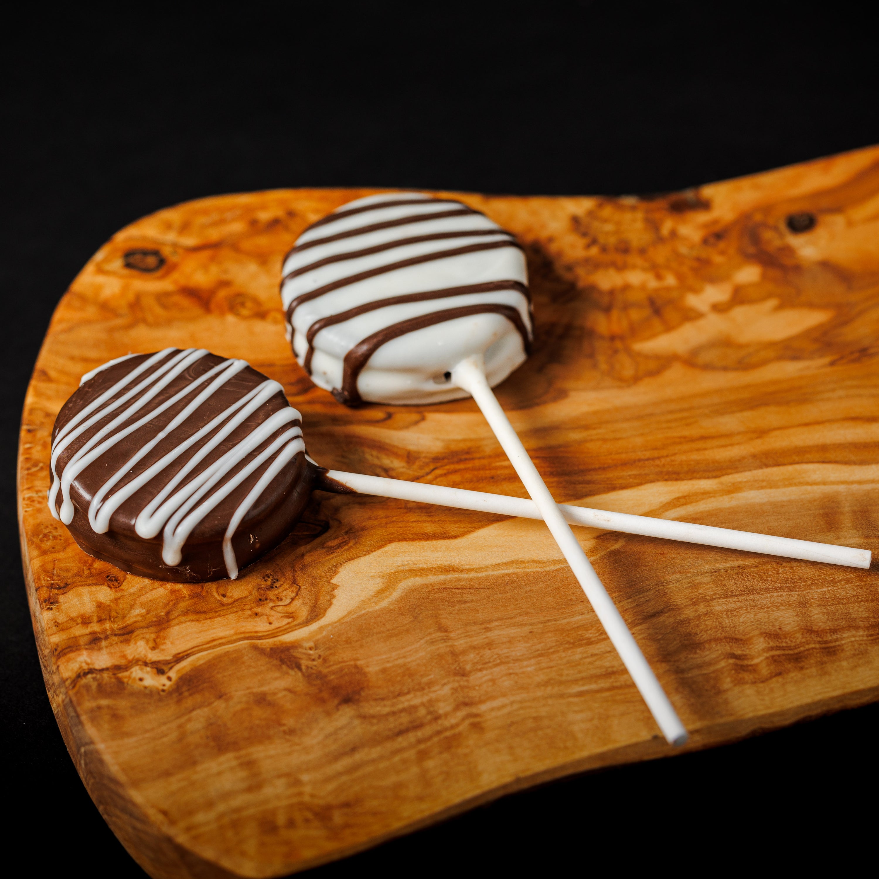 Two chocolate sandwich cookies on sticks, with white and dark chocolate drizzled on top, displayed on a wooden board.